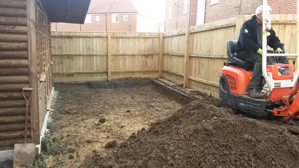 Mini excavator levelling soil beside a garden shed
