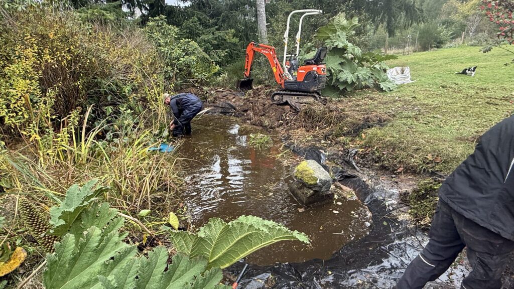 Team member clearing vegetation from the pond beside a mini excavator in Aboyne.