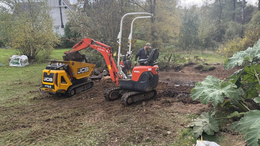 Mini excavator loading soil into a tracked dumper beside the cleared pond basin.
