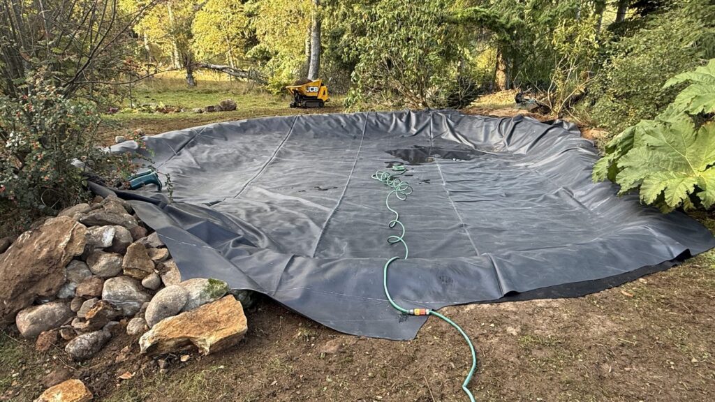 Hose laid across the new pond liner as refilling begins, with edging stones set aside.